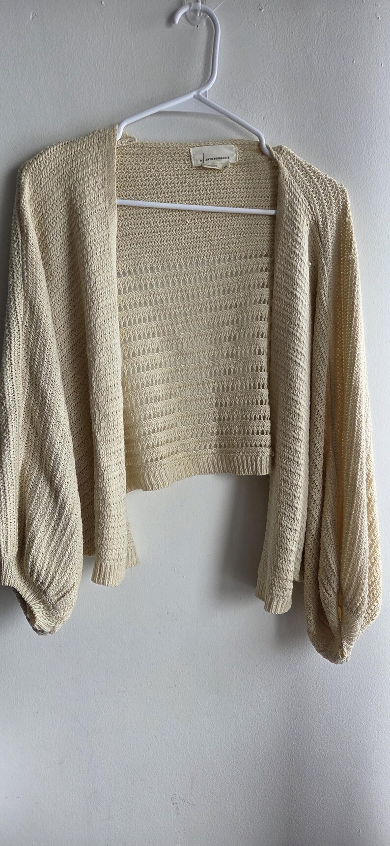 May include: Cream-colored open-front cardigan with long, voluminous sleeves. The knitwear features a textured pattern and is displayed on a white hanger against a white wall. The cardigan has a cropped length.
