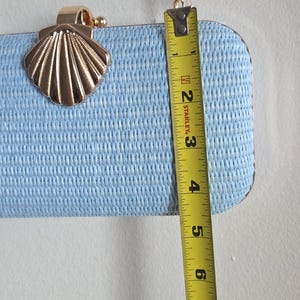 Puede incluir: Un bolso de mano rectangular de color azul claro con superficie tejida texturizada, cierre de metal dorado en forma de concha y correa de cadena dorada. Una cinta m&eacute;trica amarilla Stanley muestra una altura de aproximadamente 11,4 cm.