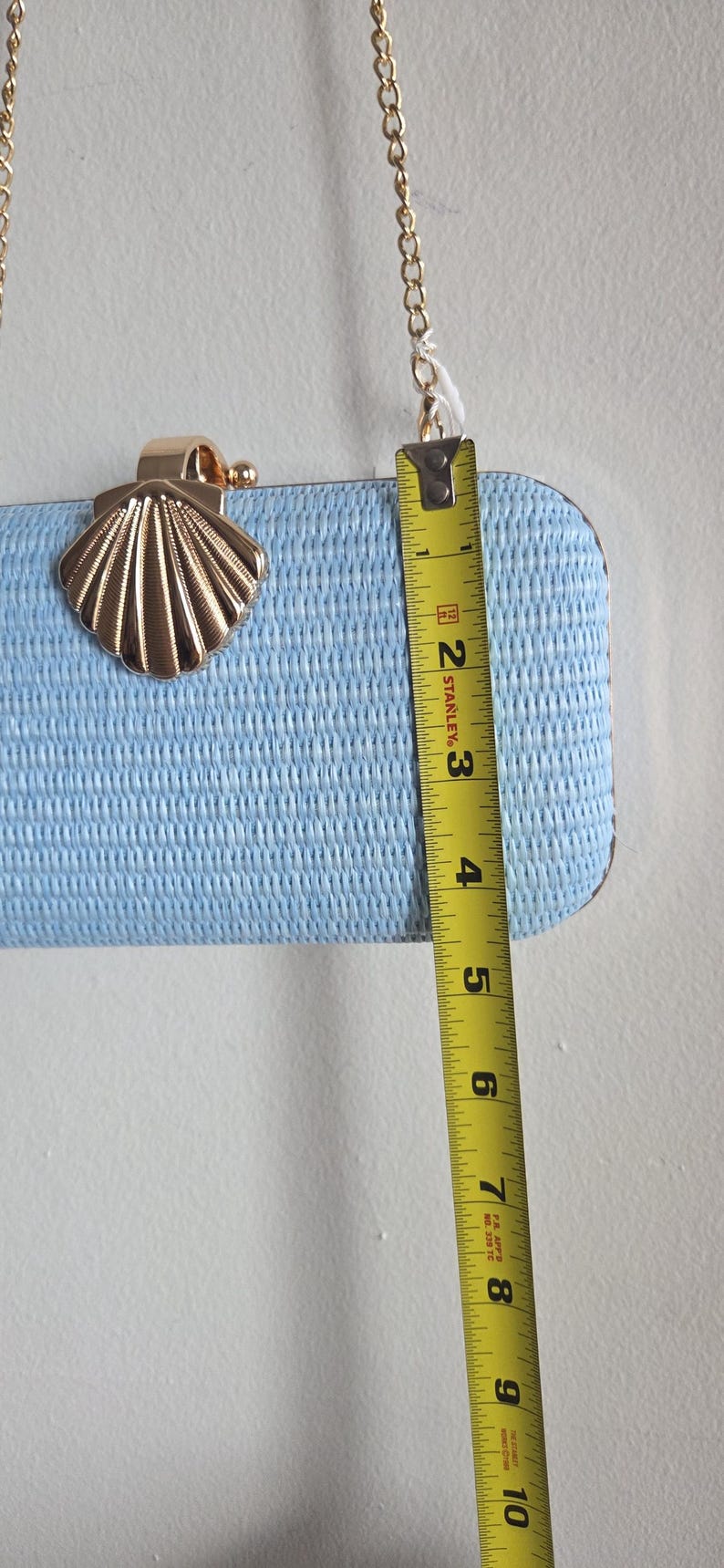 Puede incluir: Un bolso de mano rectangular de color azul claro con superficie tejida texturizada, cierre de metal dorado en forma de concha y correa de cadena dorada. Una cinta m&eacute;trica amarilla Stanley muestra una altura de aproximadamente 11,4 cm.
