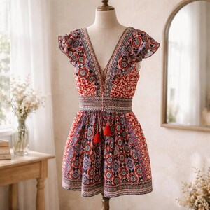 May include: A patterned mini dress with a V-neck and ruffled sleeves. The dress features a red, white, and purple floral design with a tassel tie at the waist. The dress is displayed on a mannequin.