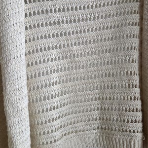 May include: Cream-colored open-front cardigan sweater. The sweater features a knitted design with a combination of solid and openwork sections. The brand label "Anthropologie" is visible at the top. The garment is hanging on a white hanger.