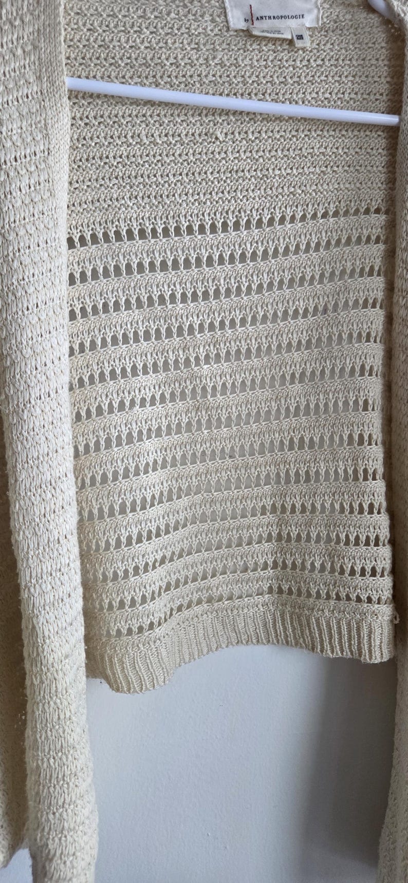 May include: Cream-colored open-front cardigan sweater. The sweater features a knitted design with a combination of solid and openwork sections. The brand label "Anthropologie" is visible at the top. The garment is hanging on a white hanger.