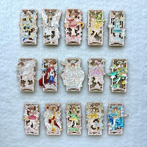 Puede incluir: Una colección de quince pines rectangulares de esmalte con varios personajes de estilo anime. Cada pin tiene un marco de filigrana dorado y representa un personaje en colores vibrantes, con detalles como alas y vestidos.