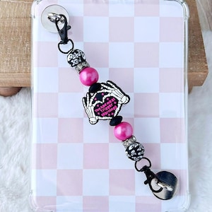 May include: A pink and black beaded phone charm with a lobster clasp. The charm features a heart-shaped design with skeleton hands and the text "You had me at Trigger Warnings". The charm also includes skull beads.