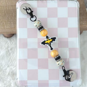 May include: A phone case with a pink and white checkered pattern. A beaded charm with black and white beads, yellow beads, and a dragon charm is attached to the case with a black clasp.