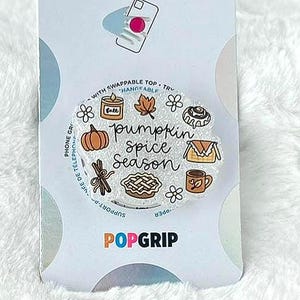 May include: A PopSockets PopGrip with a white background. The design features the words "pumpkin spice season" with illustrations of a pumpkin, pie, cinnamon sticks, and a coffee cup. The PopGrip is on a white fluffy surface.