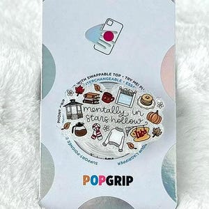 May include: A PopSockets PopGrip with a white and colorful design. The design includes the text "mentally in stars hollow" with fall-themed illustrations. The PopGrip is on a white, fluffy surface.