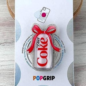 May include: A Diet Coke PopSocket phone grip on a white card. The PopSocket features a red and white Diet Coke can design with a red bow. The card has the PopSockets and PopGrip logos.