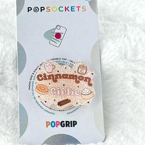 May include: A PopSockets PopGrip phone grip on a white fluffy surface. The grip features a design with the words "Cinnamon Girlie" and illustrations of a coffee cup, donut, and cinnamon roll. The PopSockets packaging is visible.