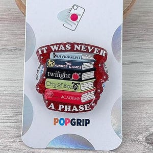 May include: A PopSockets PopGrip featuring a stack of books with the text "IT WAS NEVER A PHASE." The books include titles like "Twilight" and "The Hunger Games." The PopGrip is on a white card with the PopSockets logo.