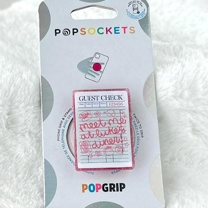 May include: A pink PopSocket phone grip featuring a miniature guest check design with handwritten notes. The PopSocket is attached to a white backing card with the PopSockets logo. The background includes a white fluffy surface and decorative items.