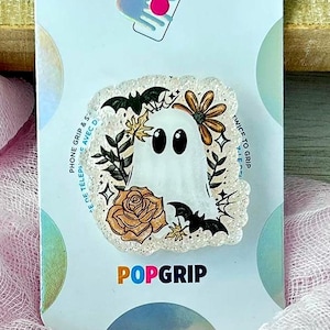 May include: A PopSockets PopGrip featuring a white ghost design with black bats, flowers, and a rose. The PopGrip is on a white card with the PopSockets logo. The background includes pink fabric and a wooden surface.