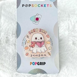 May include: A PopSockets PopGrip featuring a cartoon ghost reading a book, wearing a scarf, and surrounded by fall-themed illustrations. The text on the PopGrip reads "Cozy Girl Season". The PopGrip is on a white fluffy surface.