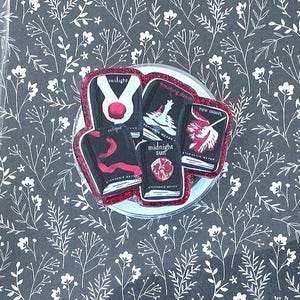 May include: A clear tablet case featuring a black background with a white floral pattern. Attached to the center is a circular grip decorated with miniature book covers from the Twilight series, including Twilight, New Moon, Eclipse, Breaking Dawn, and Midnight Sun.