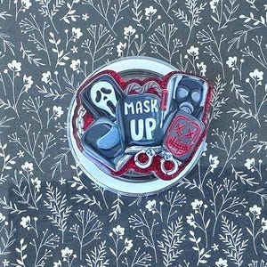 May include: A rectangular tablet case with a black background featuring a white floral pattern. Attached to the center is a circular phone grip with a red glitter border and a graphic design of horror masks and the text MASK UP in white letters.