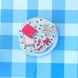 May include: A clear tablet case featuring a blue and white gingham pattern with a circular phone grip attached to the center. The grip displays a glittery background with illustrations of two books, a pair of red high heels, a red bow, and the text GOOD GIRL.