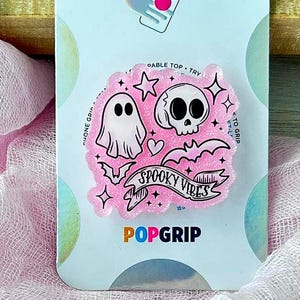 May include: A pink PopSockets PopGrip phone accessory on a retail display card. The circular grip features a graphic design with a white ghost, a white skull, small bats, stars, and a banner with the text Spooky Vibes in black lettering.