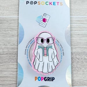 May include: A PopSockets PopGrip featuring a cartoon ghost wearing glasses and reading a book. The PopGrip is white with pink and black accents, and is attached to a light blue and white backing card. The PopGrip is designed to be used as a phone grip and stand.