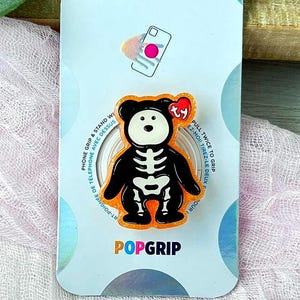 May include: A PopSockets PopGrip phone accessory featuring a black and white skeleton bear design with an orange border and a small red Ty heart tag. The product is mounted on its original light blue and white branded cardboard packaging.