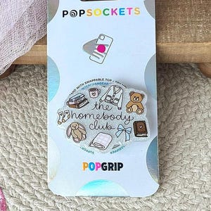 May include: A PopSockets PopGrip phone accessory on a white backing card. The circular grip features a glittery finish with the text 'the homebody club' surrounded by small illustrations of books, a mug, a cardigan, a teddy bear, socks, a blanket, and a bow.