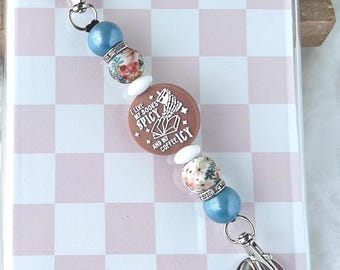 May include: A clear phone case with a pink and white checkered pattern. A beaded charm with blue, floral, and white beads is attached. The charm has a brown circular pendant with the text "I love my books spicy and my coffee icy."