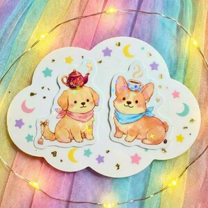 Könnte beinhalten: Zwei niedliche Hundeaufkleber auf einem wolkenförmigen Hintergrund. Einer ist ein Golden Retriever mit einer Teekanne auf dem Kopf, der andere ein Corgi mit einer Teetasse. Beide tragen Bandanas. Der Hintergrund hat Sterne, Monde und goldene Akzente.