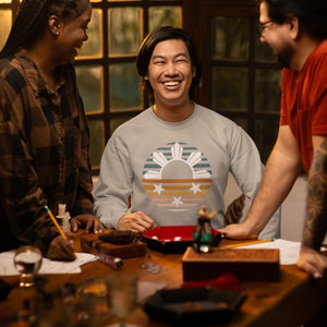 May include: Three people are sitting at a table playing a game. The person in the middle is wearing a grey sweatshirt with a retro-style design of the Philippine flag. The design features a sun with rays, a star, and stripes in shades of blue, orange, and yellow.