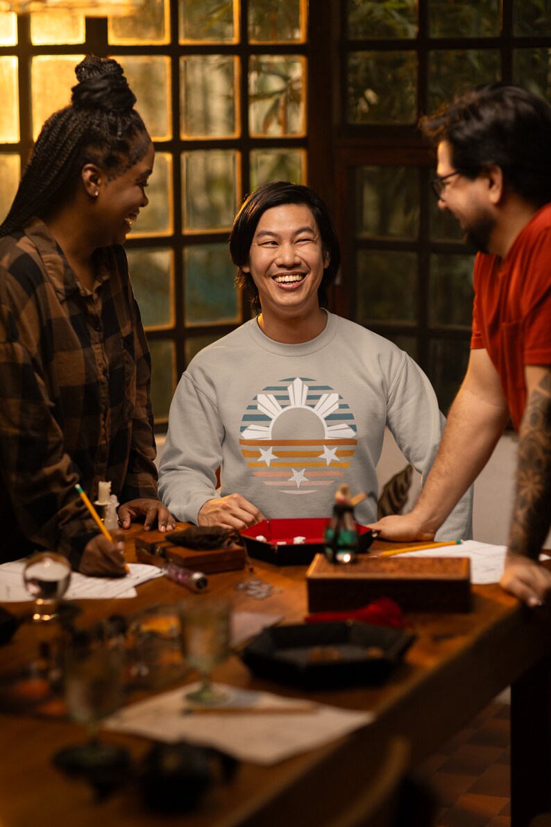 May include: Three people are sitting at a table playing a game. The person in the middle is wearing a grey sweatshirt with a retro-style design of the Philippine flag. The design features a sun with rays, a star, and stripes in shades of blue, orange, and yellow.