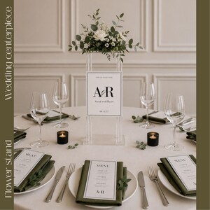May include: A clear acrylic tall flower stand centerpiece featuring a white sign with the text A&R, Anna and Ryan, and 08.12.27. The stand is topped with white roses and eucalyptus greenery, surrounded by wine glasses, menus, and black votive candles.