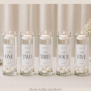 May include: Five clear cylindrical glass vases filled with water, white stones at the base, and a floating white candle on top. Each vase features a rectangular label with the text Samantha & John, Table, the number, and 08.12.28.