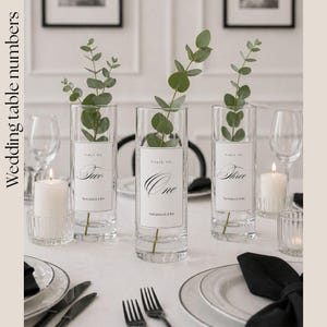 May include: Elegant wedding table setting with three clear glass vases holding eucalyptus branches. Each vase has a label with a table number: One, Two, and Three. White candles and place settings complete the sophisticated look.