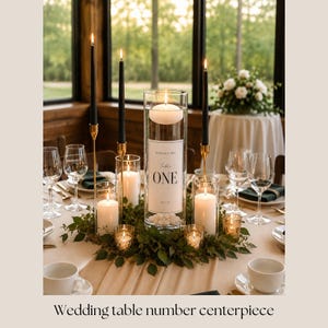 May include: A tall cylindrical glass vase centerpiece featuring a white label with the text Table ONE, Samantha & John, and a date. The arrangement includes a floating white candle, three black taper candles in gold holders, and white pillar candles.