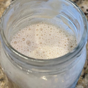 May include: A glass jar filled with a white, bubbly liquid. The liquid has a light brown hue and is filled with small bubbles.