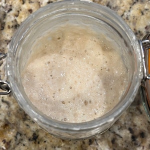 May include: A glass jar filled with a bubbly, light brown sourdough starter. The starter is active and has a foamy texture.