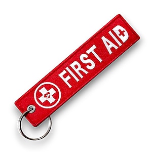 Könnte beinhalten: Ein roter, rechteckiger Schlüsselanhänger mit dem weißen Schriftzug "FIRST AID" und einem weißen Kreuzsymbol. Er hat einen silbernen Metallring. Der Hintergrund ist weiß.