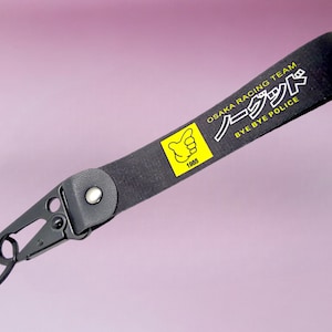 Peut inclure: Porte-clés noir avec une sangle noire et un crochet en métal argenté. La sangle porte un texte blanc qui dit "OSAKA RACING TEAM" et "BYE BYE POLICE" en japonais. Un carré jaune avec un symbole de pouce levé et l'année 1983 est sur la sangle.