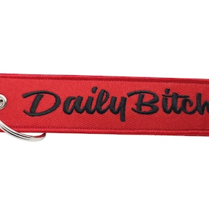 Puede incluir: Llavero de tela rojo con una frase bordada en negro "Daily Bitch".