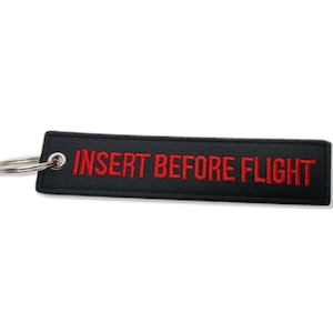 Puede incluir: Llavero de tela negro con un anillo de llave plateado. El llavero tiene un texto bordado en rojo que dice "INSERT BEFORE FLIGHT".