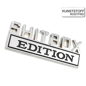 May include: Silver metal emblem with the words "Shitbox Edition" in black lettering. The emblem is rectangular with a raised border.