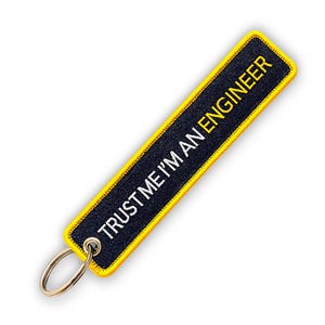 Könnte beinhalten: Gestickter Schlüsselanhänger mit dem Text "TRUST ME I'M AN ENGINEER" in Weiß und "ENGINEER" in Gelb auf schwarzem Hintergrund. Der Schlüsselanhänger hat einen gelben Rand und einen silbernen Schlüsselring.