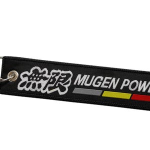 Op de afbeelding: Zwarte sleutelhanger met een wit geborduurd logo met de tekst "Mugen Power" met Japanse karakters erboven. De sleutelhanger heeft een streepontwerp in rood, geel en grijs.