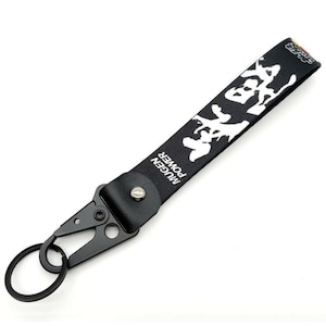 May include: Black keychain strap featuring white Japanese characters and the text "MUGEN POWER". It's connected to a black metal carabiner clip and a split ring. The background is white.