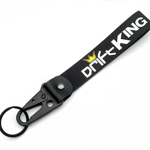 Puede incluir: Llavero negro con un logotipo "DRIFT KING" en blanco y negro y una corona dorada. El llavero tiene un clip de metal y un anillo.