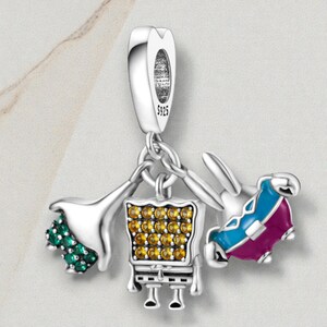 May include: A silver charm with three dangling pendants. One pendant is a green and silver flower, another is a yellow and silver chair, and the third is a blue and purple rabbit. The charm has a loop at the top for attaching to a bracelet or necklace.