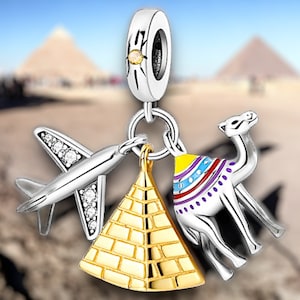 May include: Silver charm with a travel theme, featuring a detailed airplane with clear gem accents, a gold pyramid, and a silver camel with colorful saddle details. The charm is designed to be attached to a bracelet or necklace.