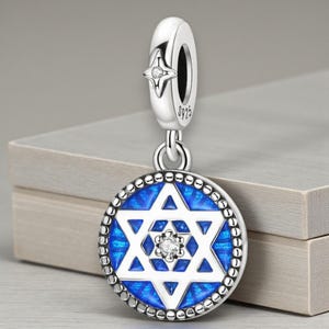 May include: A silver charm with a Star of David design. The charm has a blue enamel background with a white Star of David and a small clear gem in the centre. The top has a star-shaped accent with a small gem.