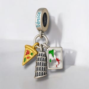 Può includere: Un charm d'argento con la scritta "Venice" in lettere turchesi. Il charm presenta tre ciondoli: una fetta di pizza, la Torre di Pisa e una mappa dell'Italia. La fetta di pizza è dorata con dettagli rossi e verdi.