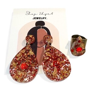 May include: A set of resin jewelry, including teardrop earrings and a ring. The earrings and ring feature red glitter and gold leaf inclusions. The earrings have a post back. The ring is a wide band. The jewelry is displayed with a card that says "Shop 2dye 4 JEWELRY."