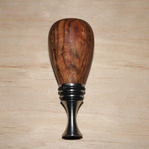 May include: A wooden bottle stopper with a dark brown, teardrop-shaped top. The stopper has a silver-colored metal base with a decorative design. The wood grain is visible, showing a natural pattern. The stopper is designed to fit into a bottle.