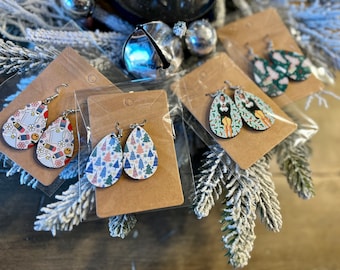 Puede incluir: Tres pares de pendientes colgantes con diferentes diseños. El primer par presenta un fondo blanco con útiles escolares coloridos, el segundo par tiene un fondo blanco con árboles de Navidad azules y rosas, y el tercer par tiene un fondo verde con un personaje de dibujos animados que lleva un atuendo verde y rojo.